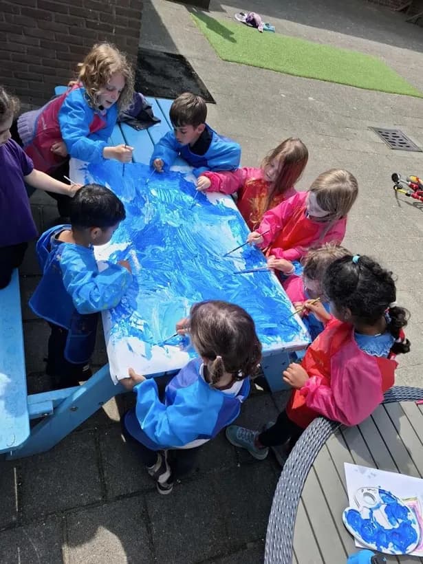 Children in colorful smocks painting a large mural together outdoors